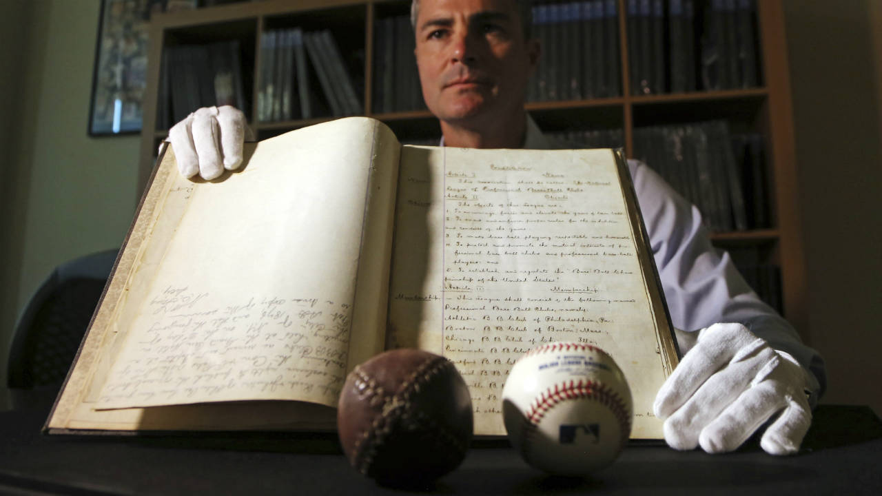 This-May-18,-2017,-photo-shows-Dan-Imler,-vice-president-of-SCP-Auctions,-with-the-bound-volume-of-the-1876-constitution-that-founded-the-National-League-of-Professional-Base-Ball-and-the-modern-business-of-big-league-sports,-that-is-going-up-for-sale-at-SCP-Auctions-in-Laguna-Niguel,-Calif.-A-replica-of-a-period-baseball-is-at-left;-a-modern-ball-is-at-right.-SCP-Auctions-expects-the-papers-to-draw-millions-when-the-auction-starts-Wednesday,-May-24.-(Reed-Saxon/AP)