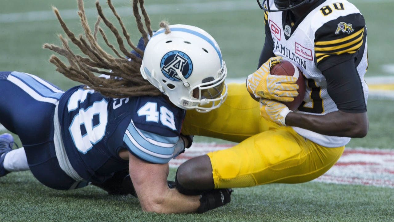 The-hair-flies-from-beneath-the-helmet-of-Toronto-Argonauts-linebacker-Bear-Woods-(48)-as-he-tackles-Hamilton-Tiger-Cats-wide-receiver-Terrence-Toliver-during-the-first-half-of-CFL-exhibition-football-action-in-Hamilton-on-Friday,-June-16,-2017.-(Peter-Power/CP)