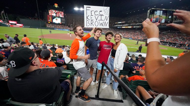 Kevin Tehansky, left, holds a sign in support of broadcaster Kevin Brown while photo bombing a group of people posing for pictures at Oriole Park at Camden Yards in the eighth inning of a baseball game between the Baltimore Orioles and the Houston Astros, Tuesday, Aug. 8, 2023, in Baltimore. (Julio Cortez/AP)