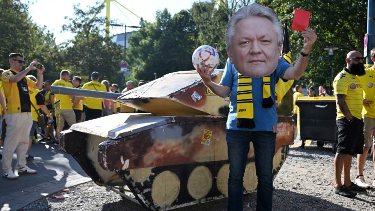 A dummy tank stands next to an activist wearing a mask with the likeness of Rheinmetall CEO Armin Papperger and holding up a red card as BVB's supporters plan to start the Bundesliga season with protests against the club’s sponsorship deal with German arms manufacturer Rheinmetall ahead of the soccer game between Borussia Dortmund and Eintracht Frankfurt, in Dortmund, Germany, Saturday Aug. 24, 2024. (Bernd Thissen/dpa via AP)