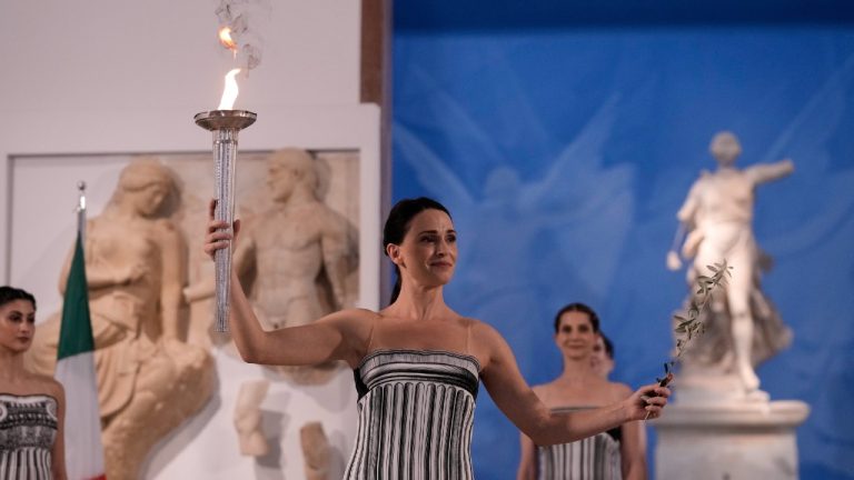 Actress Mary Mina, playing the role of high priestess, holds a torch with the Olympic flame and an olive branch during the flame lighting ceremony for the Milan Cortina 2026 Winter Olympics, at the archaeological museum of Olympia, Greece, Wednesday, Nov. 26, 2025. (Thanassis Stavrakis/AP)