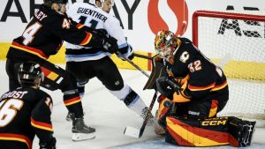 Utah Mammoth's Dylan Guenther, centre, has his shot blocked by Calgary Flames goalie Dustin Wolf, right, as Joel Hanley checks during third period NHL hockey action in Calgary on Saturday, Dec. 6, 2025. (Jeff McIntosh/CP)