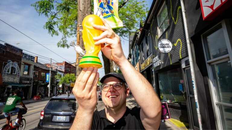 Sean Kady, owner of Cosmic Charlies, holds up a bong resembling the FIFA World Cup trophy in front of a banner promoting the tournament as he poses for a photograph, outside his shop on Queen Street West in Toronto on Thursday, June 4, 2026. (Arlyn McAdorey/CP)
