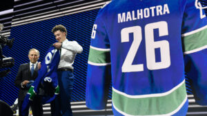 Caleb Malhotra, right, puts on a jersey after being drafted by the Vancouver Canucks as NHL Commissioner Gary Bettman, left, looks on during the NHL hockey draft Friday, June 26, 2026, in Buffalo, N.Y. (Adrian Kraus/AP)