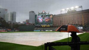 A person stands under an umbrella while it rains as a tarp covers the field ahead of a baseball game between the Baltimore Orioles and the Toronto Blue Jays, Monday, Sept. 21, 2026, in Baltimore. (Stephanie Scarbrough/AP)